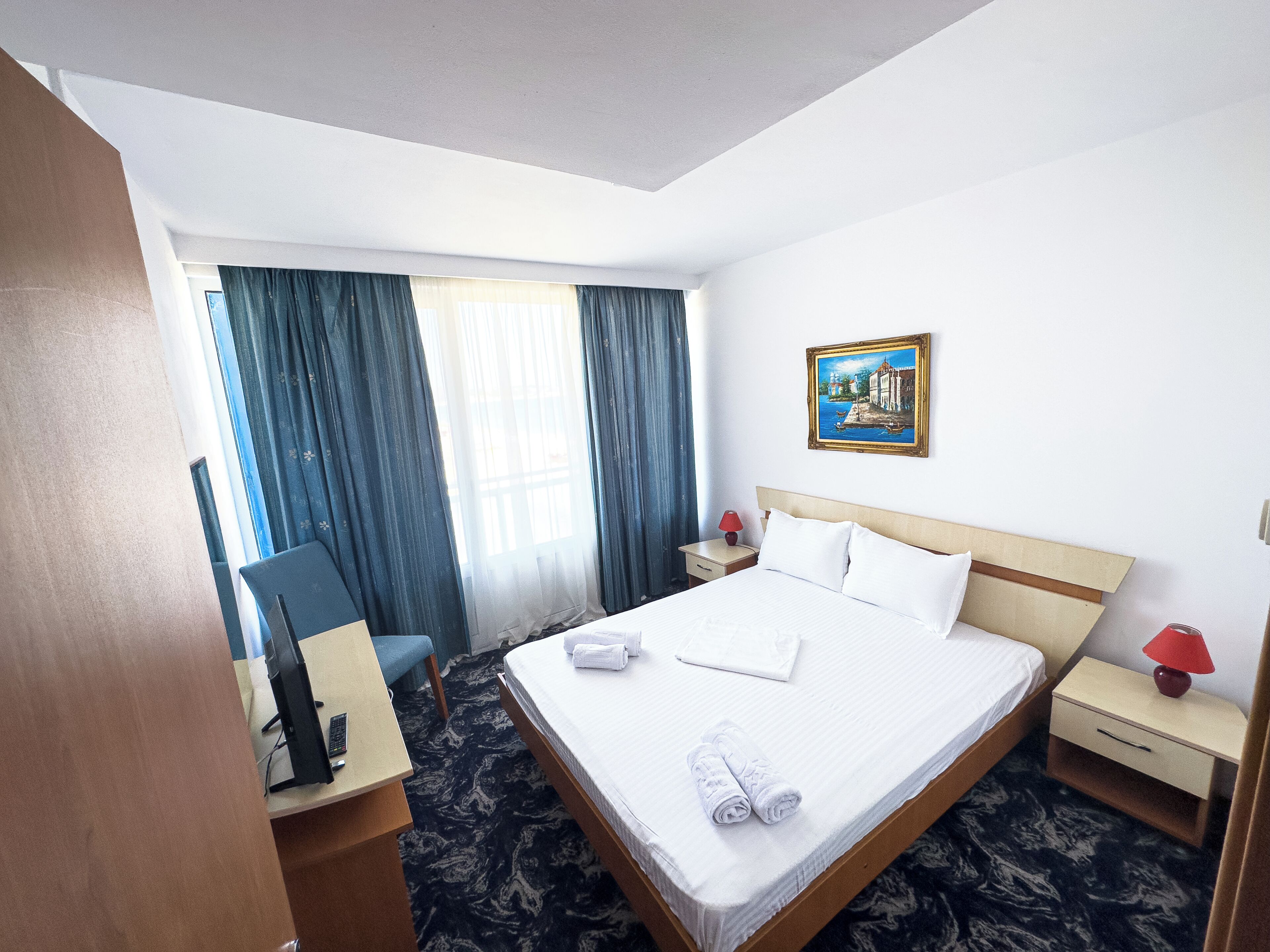 A hotel room with a bed, a desk with a computer, a chair, a nightstand with a lamp, and a painting on the wall.