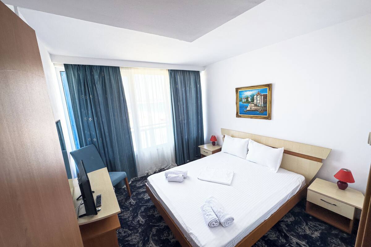 A hotel room with a bed, a desk with a computer, a chair, a nightstand with a lamp, and a painting on the wall.