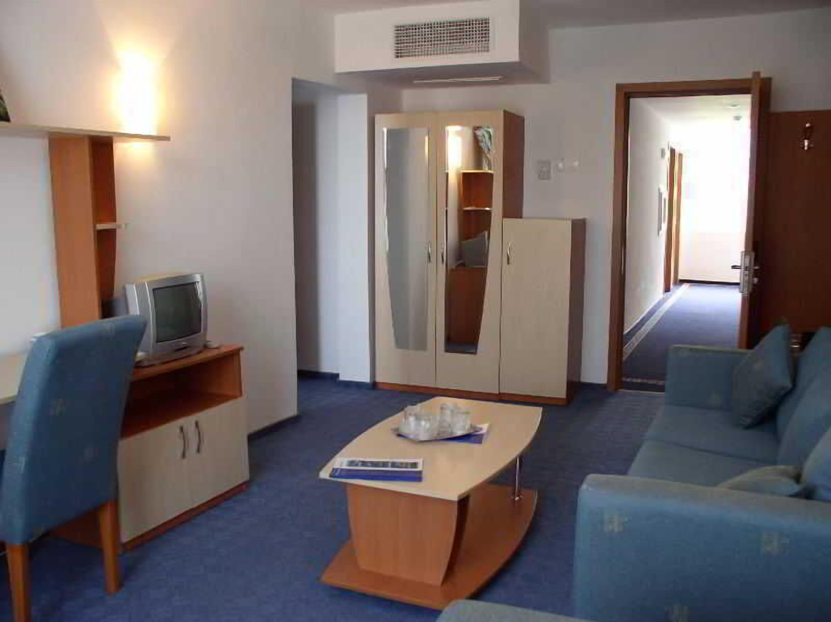 A room with a sofa, a small table, a TV on a shelf, a wardrobe, and a door leading to another room.