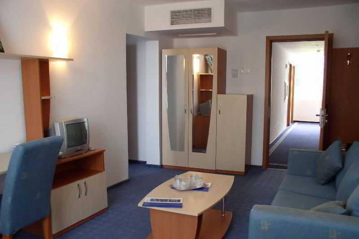 A room with a sofa, a small table, a TV on a shelf, a wardrobe, and a door leading to another room.