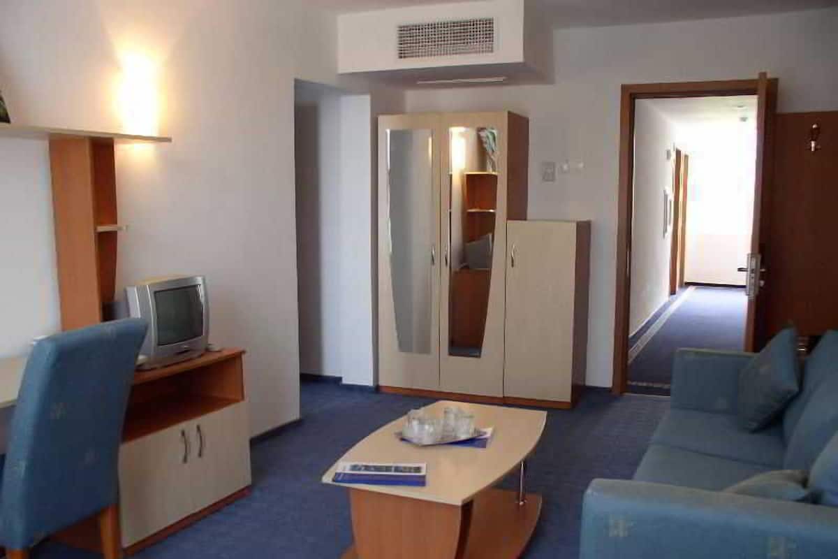 A room with a sofa, a small table, a TV on a shelf, a wardrobe, and a door leading to another room.