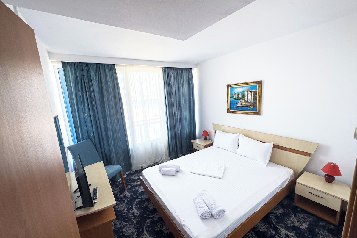 Una habitación de hotel con una cama, un escritorio con una computadora, una silla, una mesita de noche con una lámpara y un cuadro en la pared.