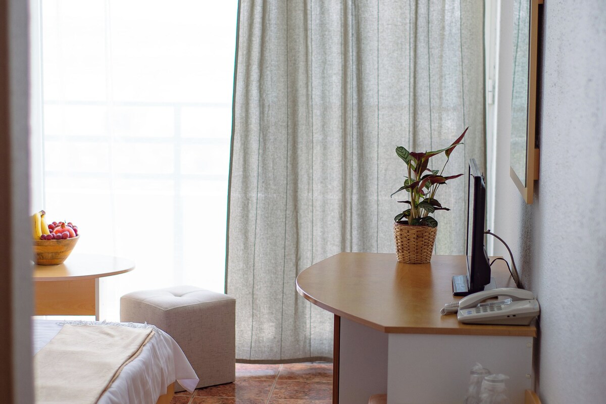 Habitación de hotel con cama, una mesita, escritorio, teléfono, espejo y ventana con cortinas.