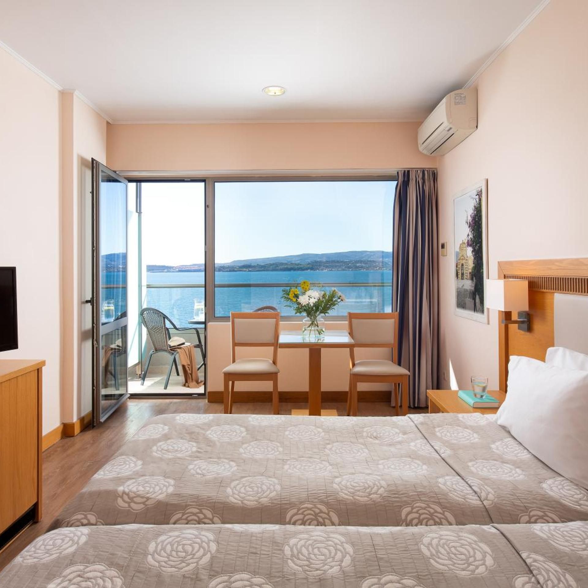 A hotel room with a bed, a desk with chairs, a view of the sea, and a painting on the wall.