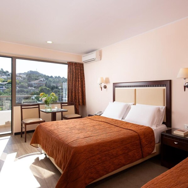 A hotel room with a large bed, a desk with a chair, a small table with a vase, and a view of a cityscape. - Mediterranee (KEFALONIA)