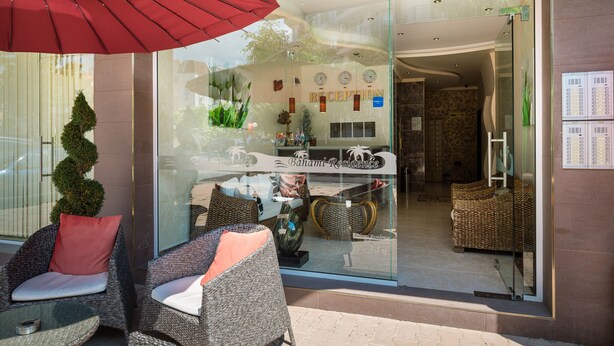 A reception area with wicker furniture, a red umbrella, and a glass wall reflecting the interior. - Hotel Bahami Residence (Sunny Beach)