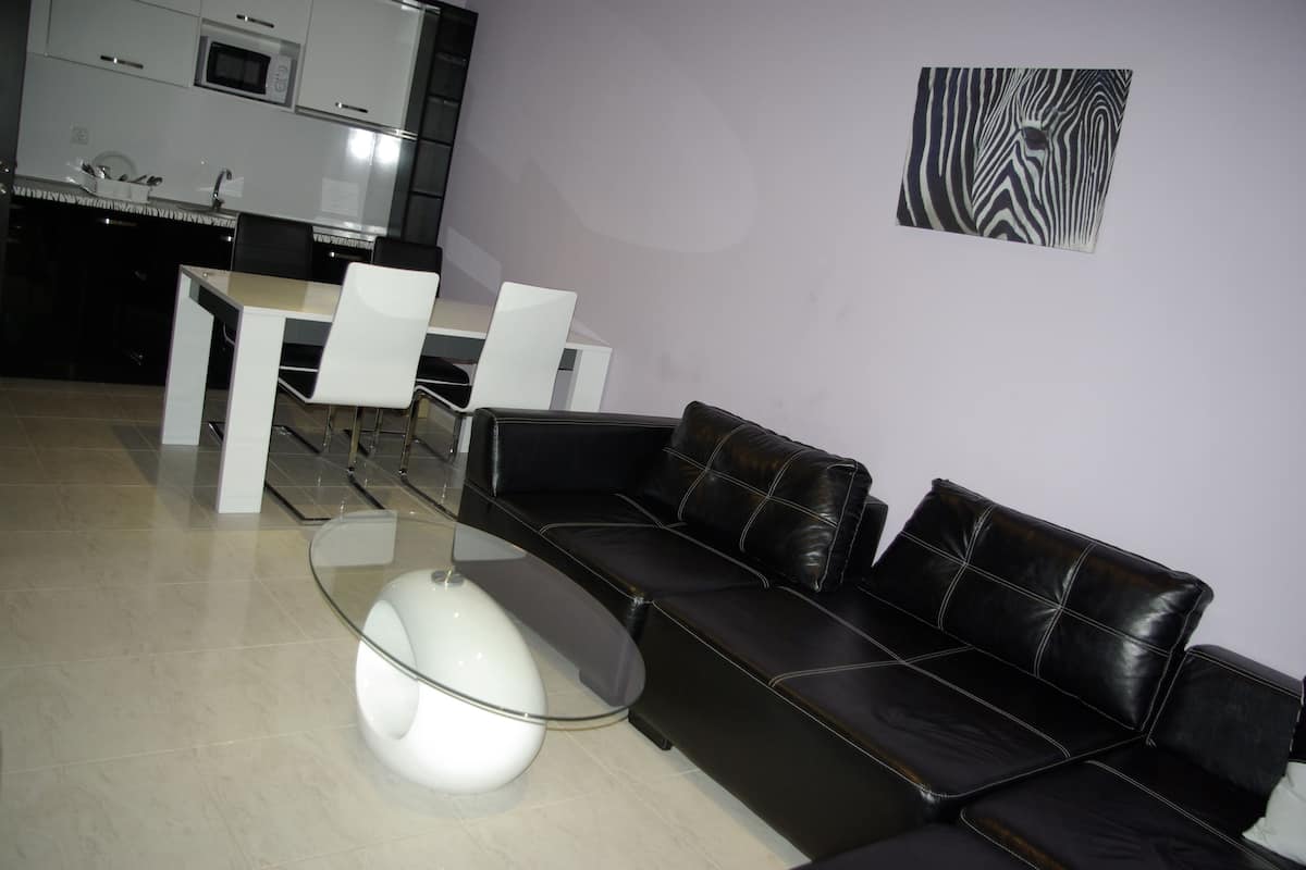 A modern living room with a black leather sofa, a glass coffee table, and a dining area with white chairs and a white table.
