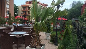 An outdoor patio area with wicker furniture, potted plants, and a red umbrella. - Hotel Bahami Residence (Sunny Beach)