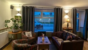 A cozy living room with a fireplace, two armchairs, a sofa, and a view of a parking lot through a window. - The Bog Roy Inn (Inverness)
