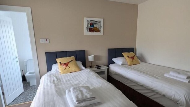 A hotel room with two single beds, a small bedside table with a lamp, a wall-mounted painting, and a door leading to the bathroom. - The Bog Roy Inn (Inverness)