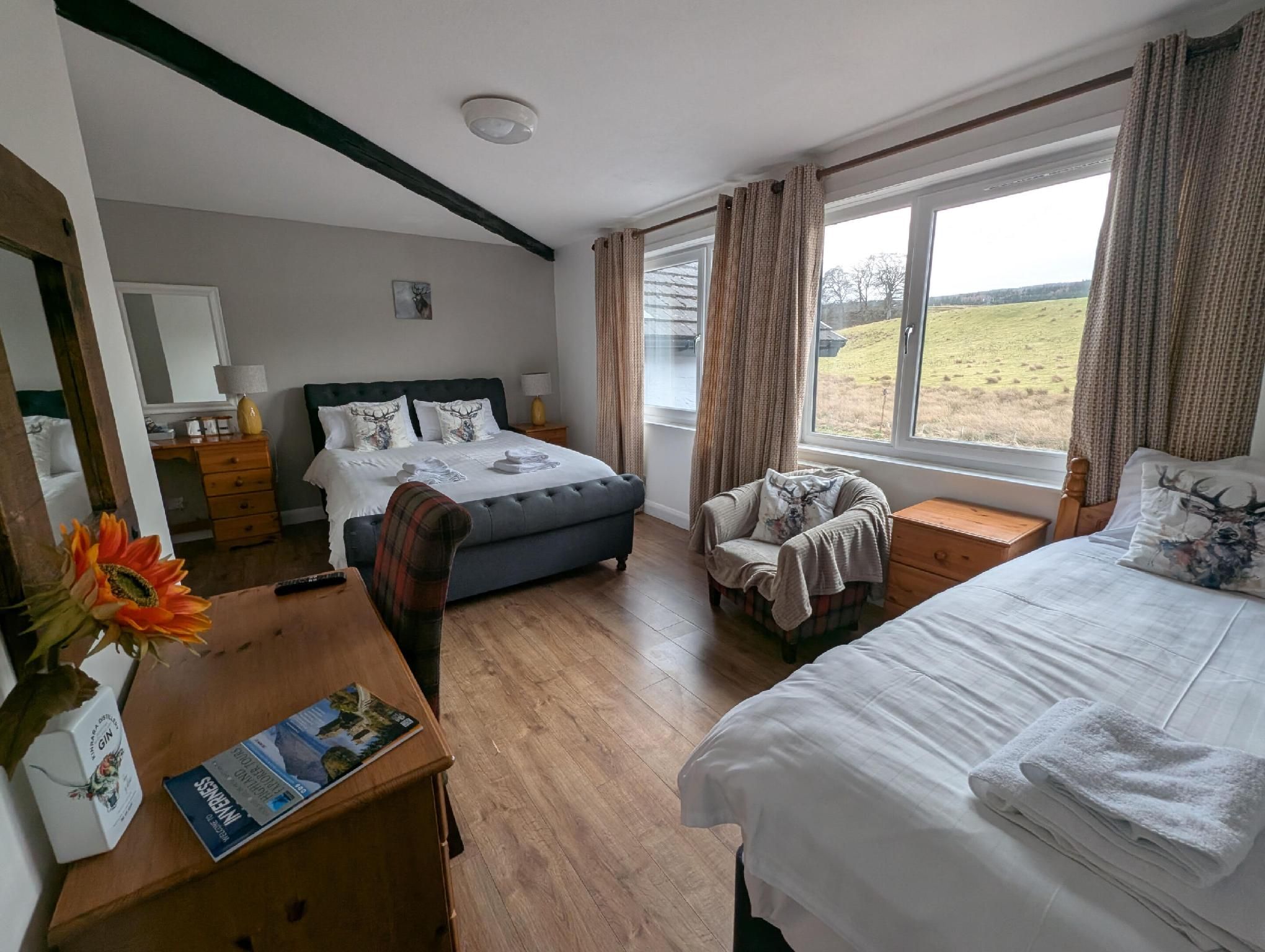 A hotel room with a large bed, a smaller bed, a sofa, a wooden table with a magazine, a vase with flowers, and a window with a view of the countryside.