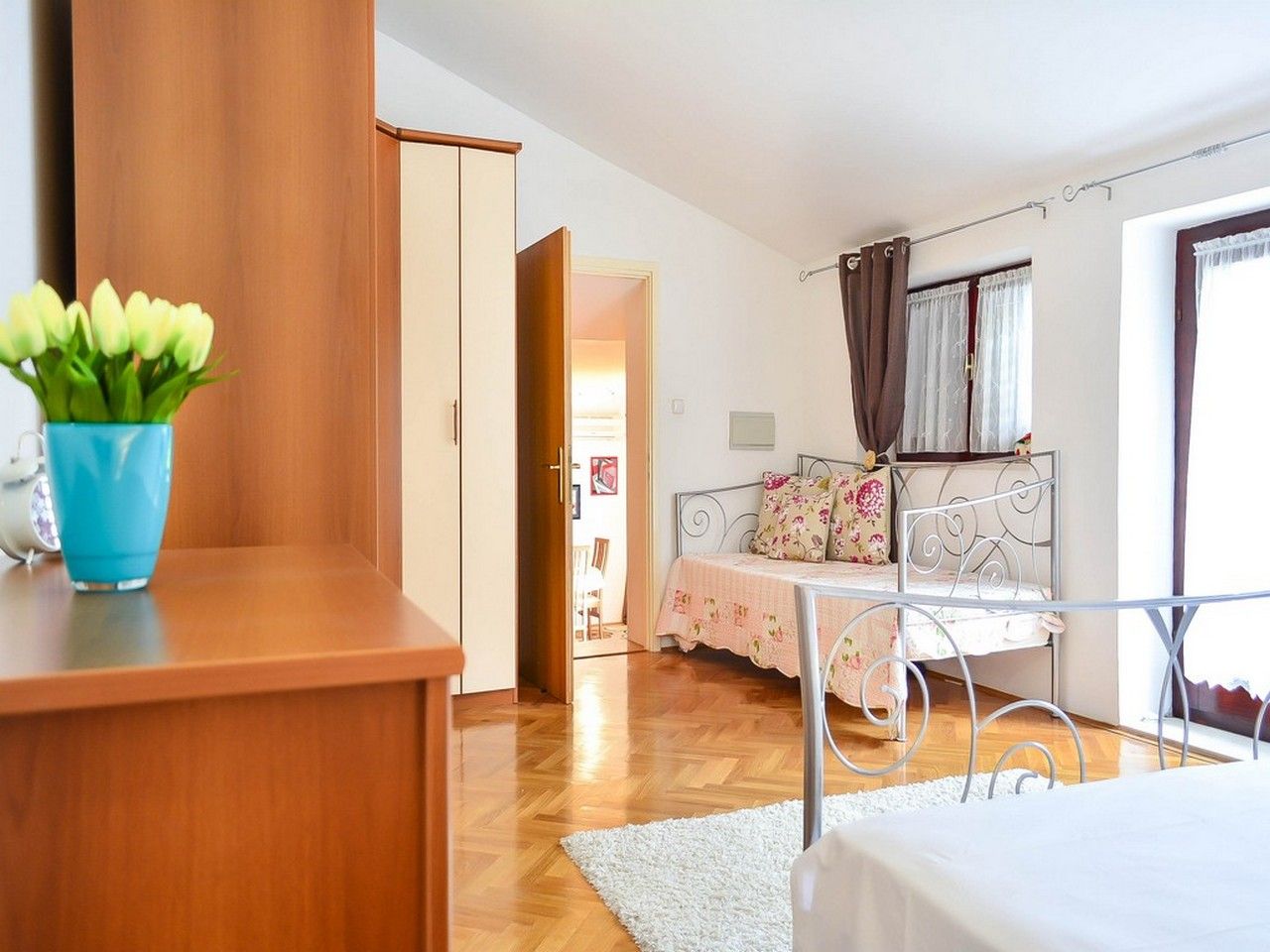 A bedroom with a bed, a bedside table, a wardrobe, a window with curtains, and a view into another room.