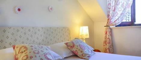 A bedroom with a bed, pillows, a nightstand, a lamp, and a window with curtains.