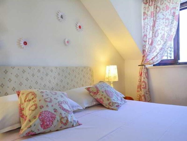 A bedroom with a bed, pillows, a nightstand, a lamp, and a window with curtains.