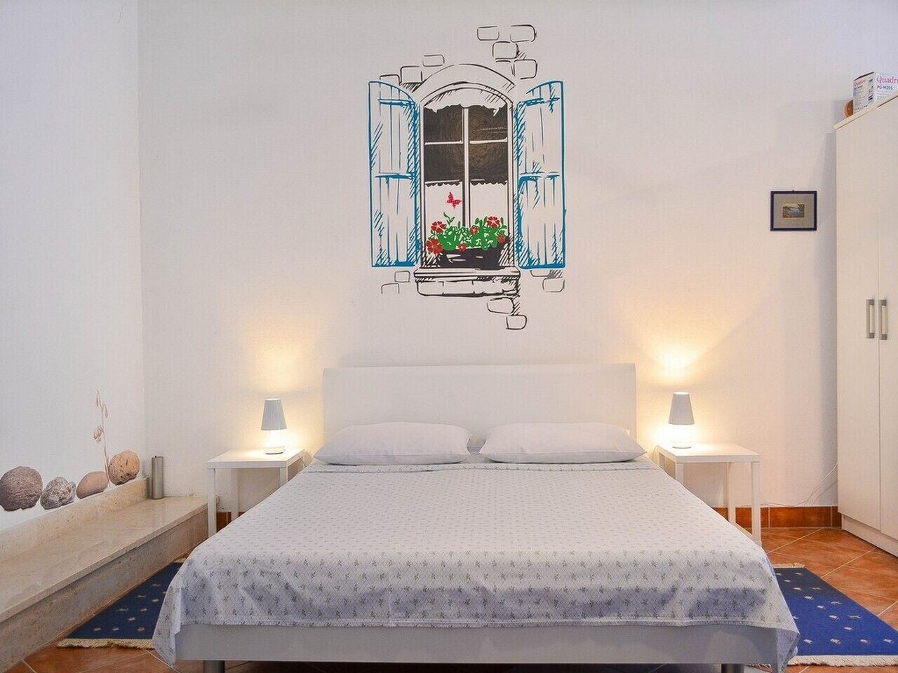 Un dormitorio con cama, dos mesitas de noche, un ventanal con vistas a flores y un armario.