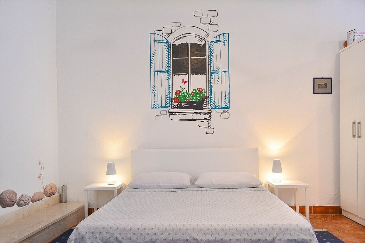 A bedroom with a bed, two bedside tables, a window with a view of flowers, and a wardrobe.