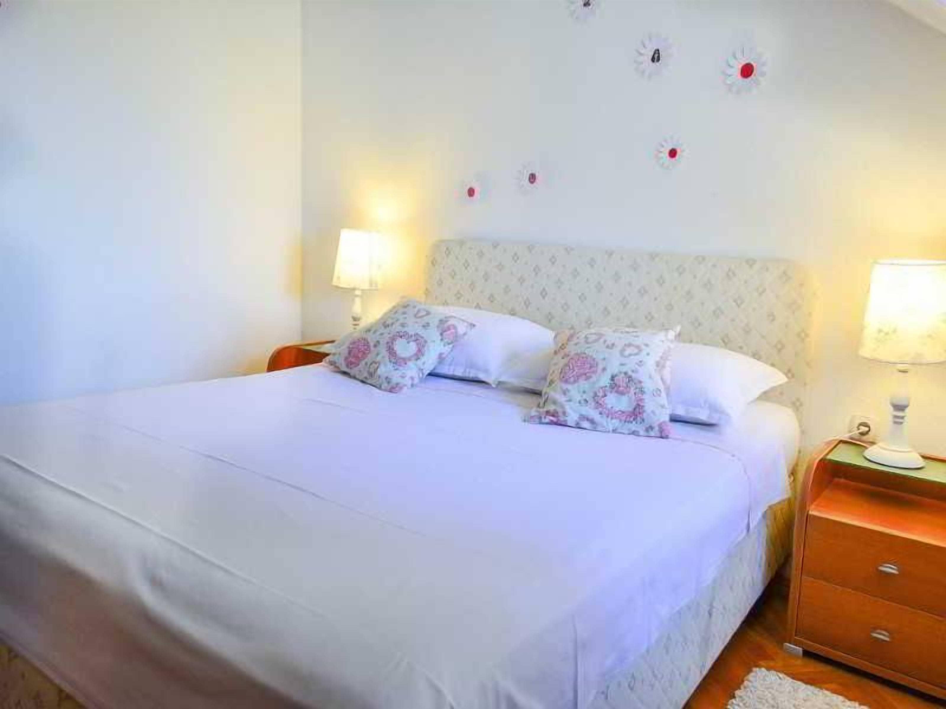 A neatly made bed with floral-patterned pillows, a bedside table with a lamp, and a wall with decorative elements.