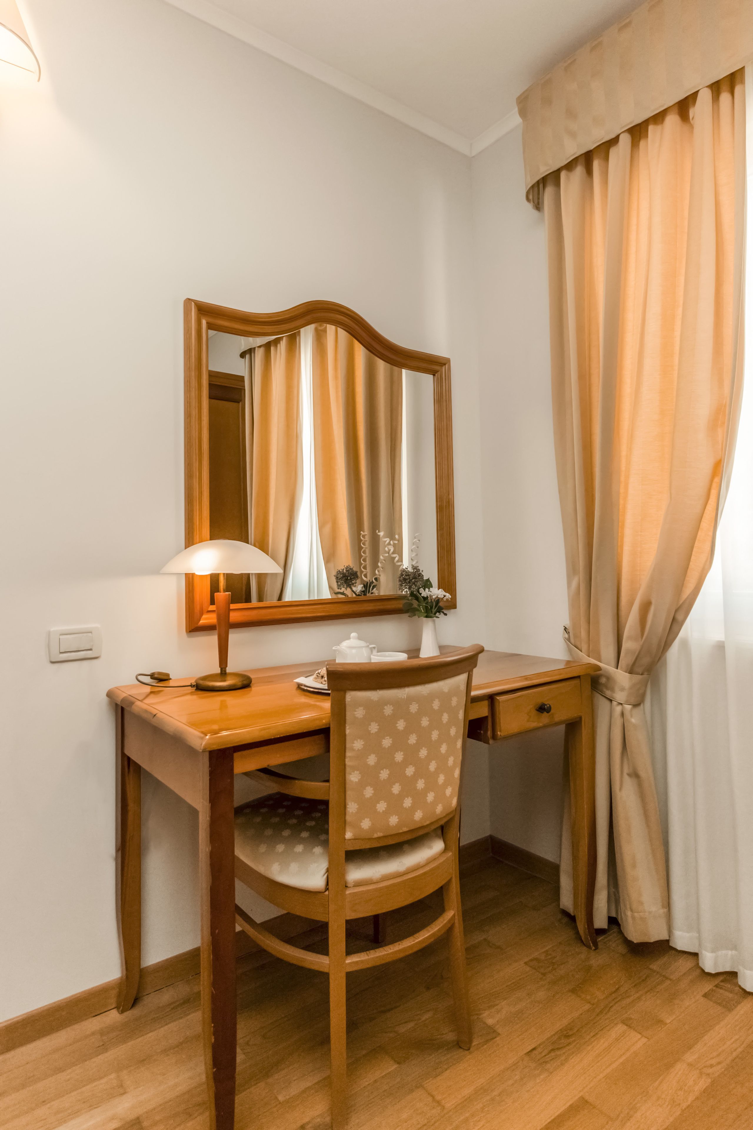 Un bureau en bois, une lampe, une chaise, un miroir et un petit vase avec des fleurs.