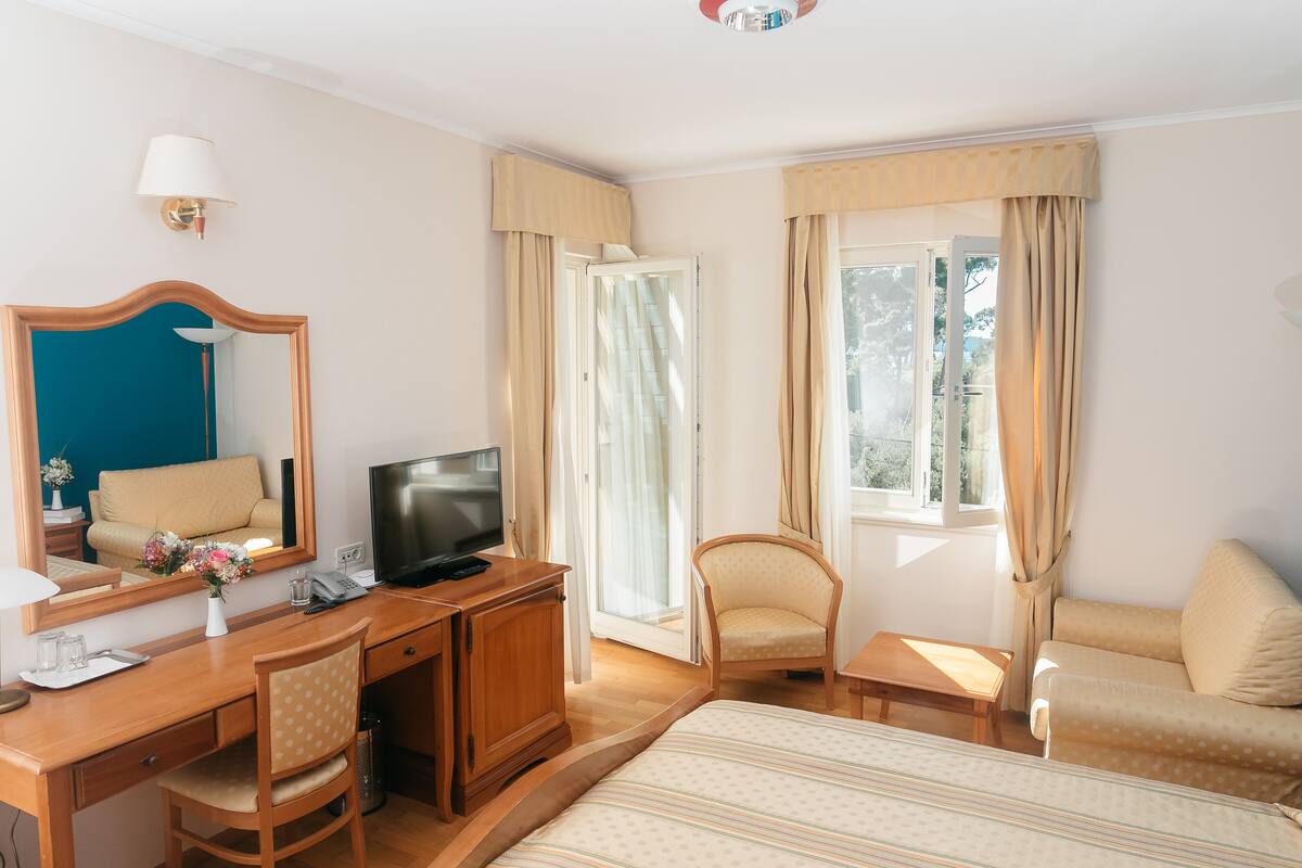 A hotel room with a bed, a desk with a TV, a chair, a sofa, and a window with curtains.