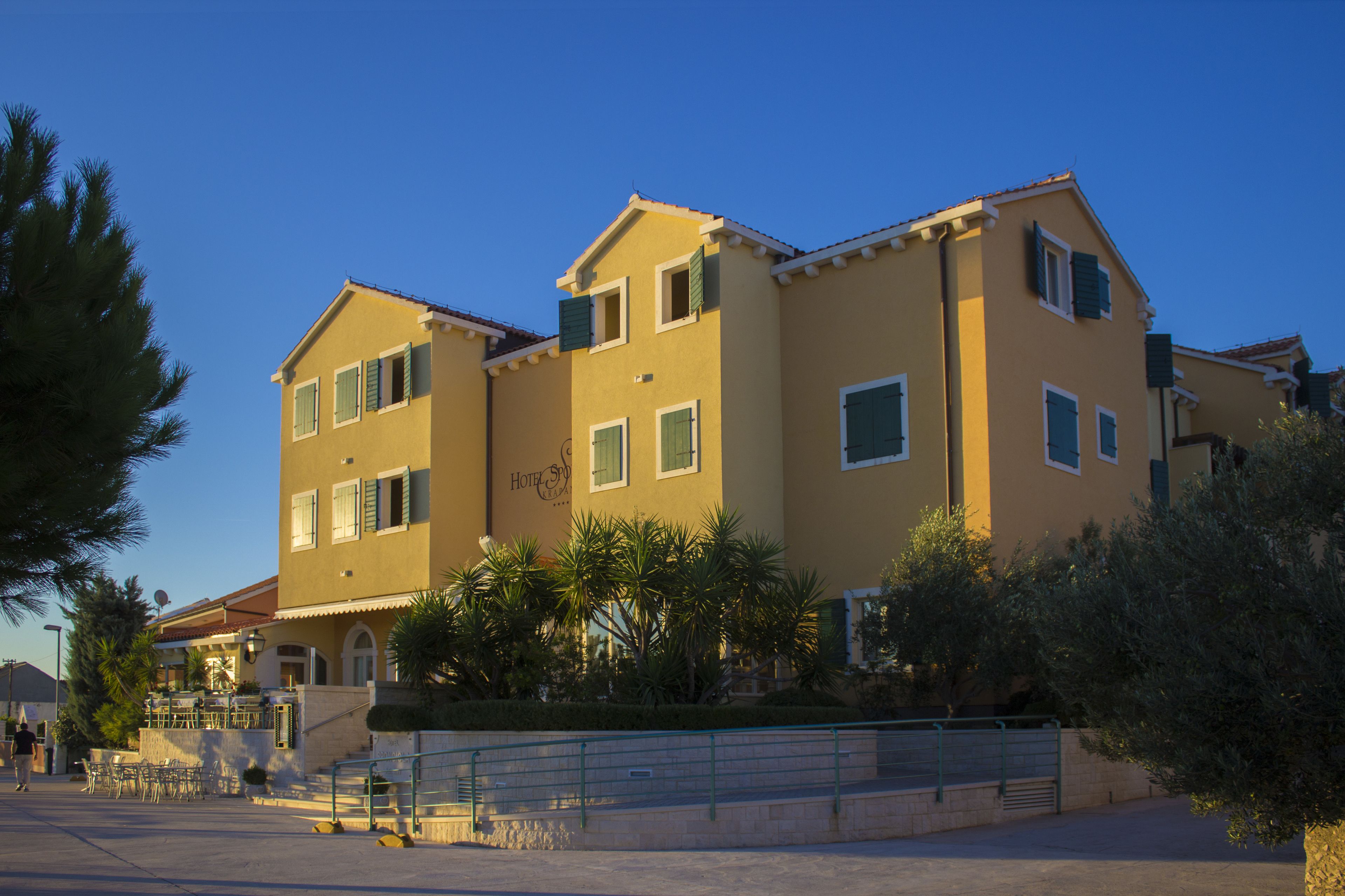 A two-story hotel with a yellow facade, multiple windows with shutters, and a visible sign indicating it is a hotel.