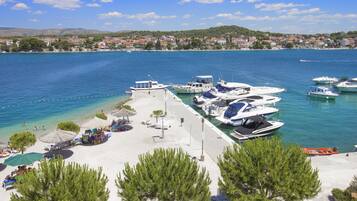 A marina with boats docked, a sandy beach area with umbrellas, and a backdrop of a coastal town.