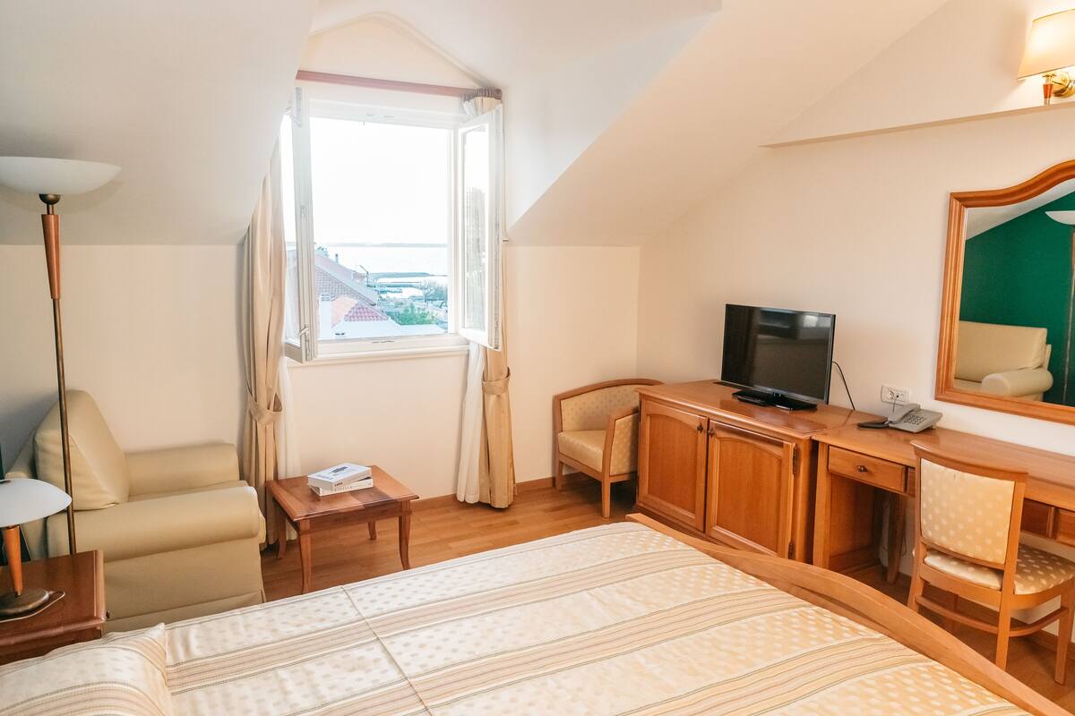A hotel room with a bed, a desk with a TV, a chair, a lamp, and a window with curtains.