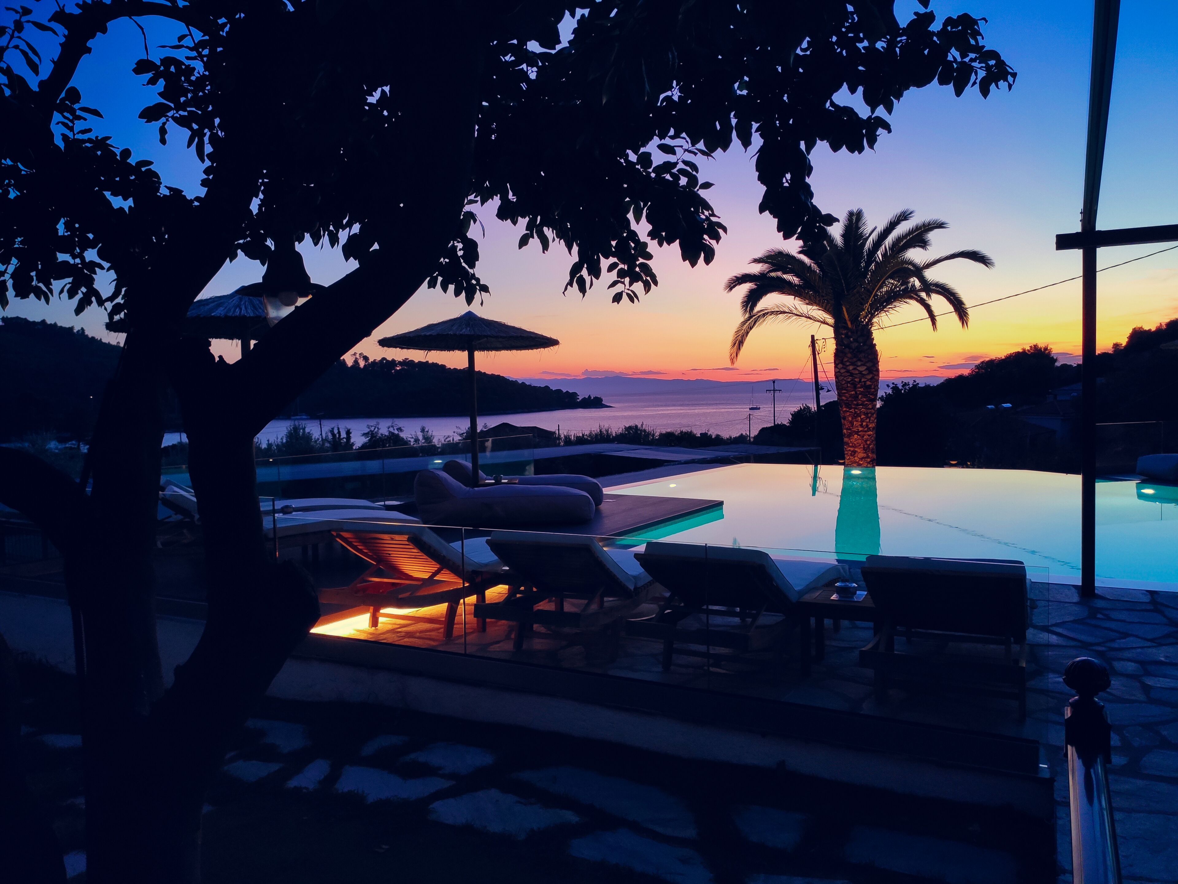 A poolside area with lounge chairs and umbrellas, overlooking a body of water during sunset.
