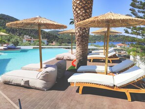 Un espace aménagé au bord de la piscine, avec des chaises longues et des parasols en chaume, entouré de palmiers et offrant une vue sur la mer.