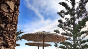 Een strandlounge met ligbedden en rieten parasols, met uitzicht op een waterpartij en een bergachtig landschap.