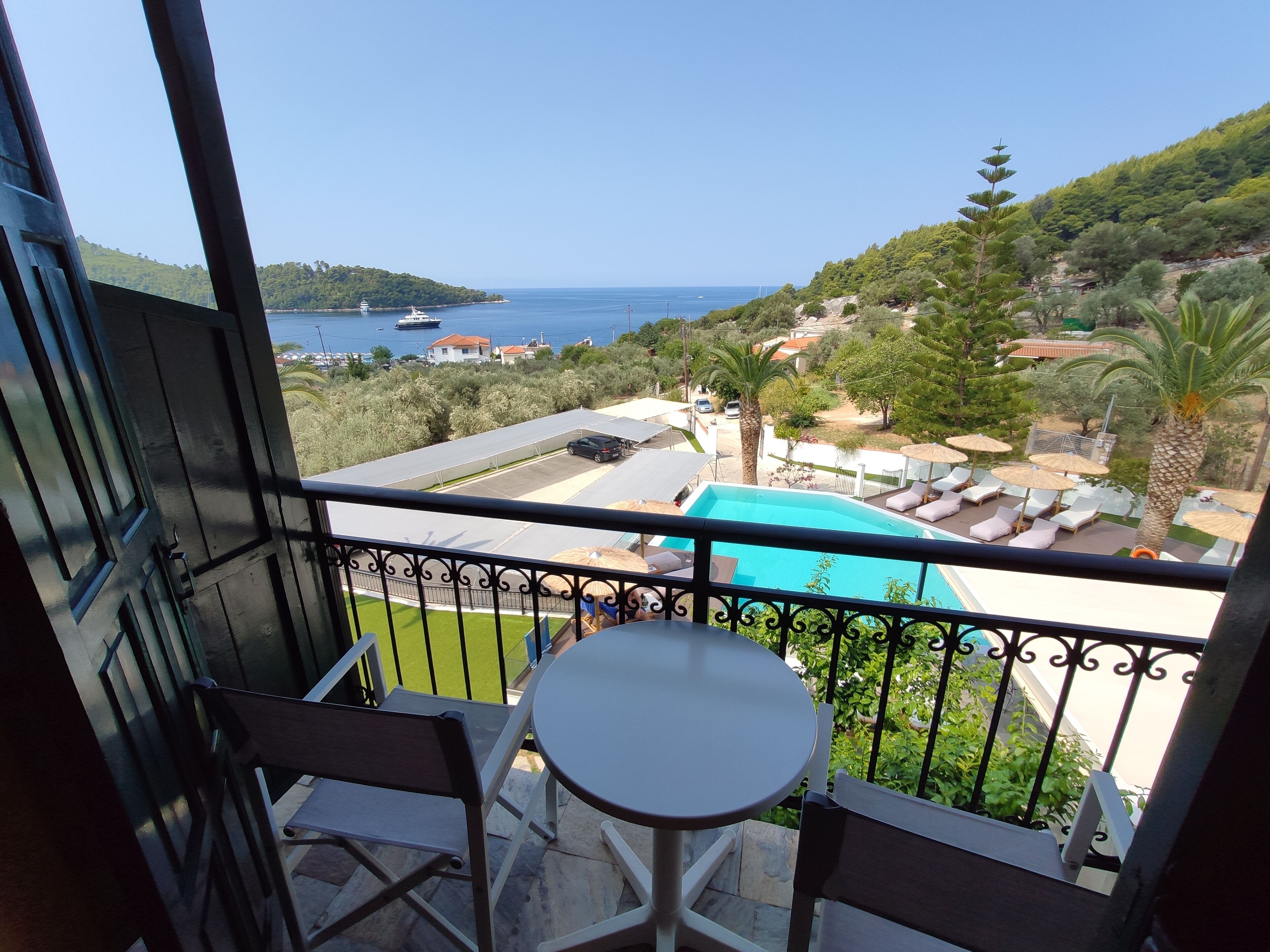 A balcony with a view of a swimming pool, lounge chairs, and a distant view of the sea and hills.
