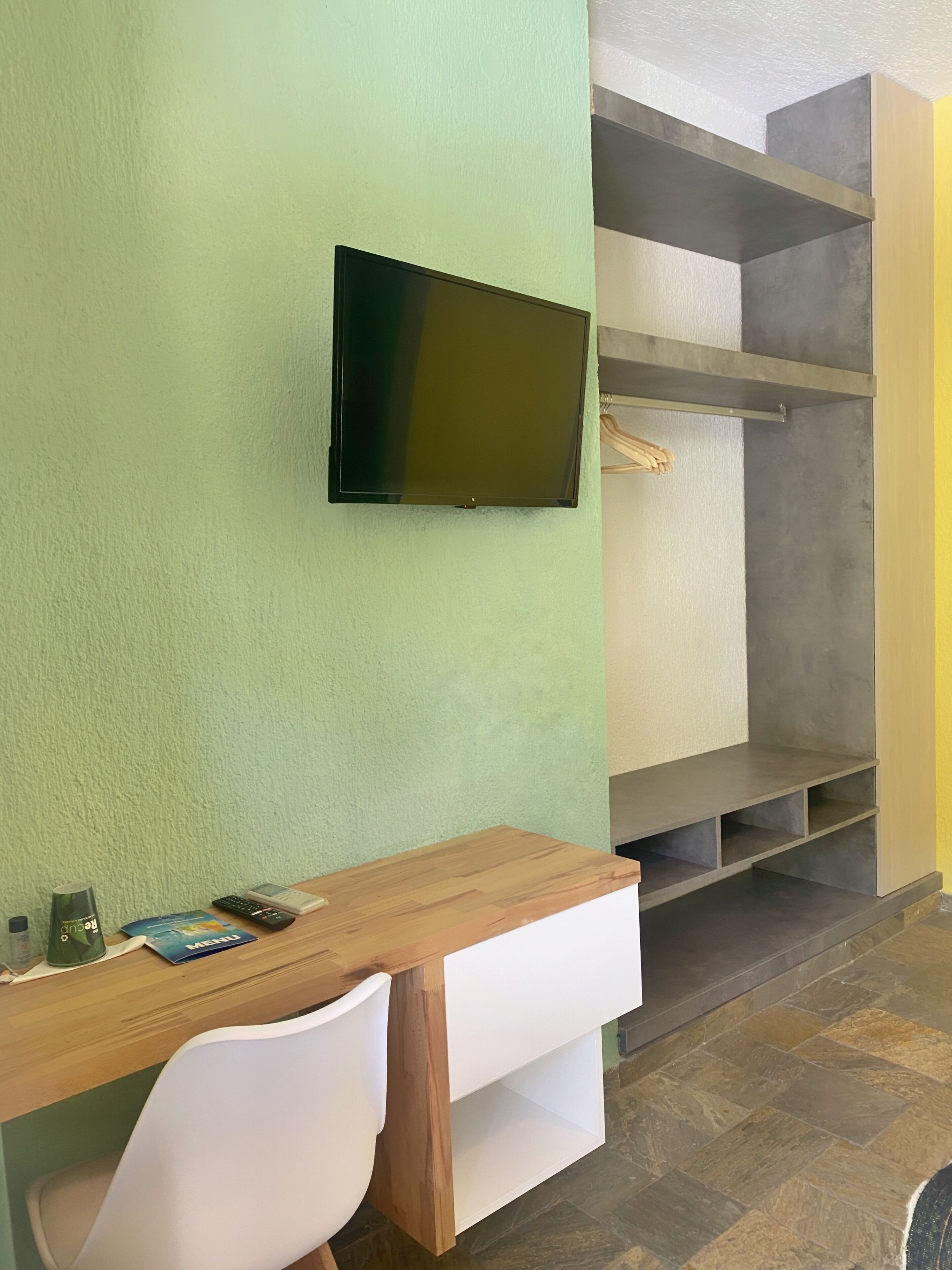 A room with a green wall, a mounted TV, a wooden desk with a chair, and a built-in shelving unit.