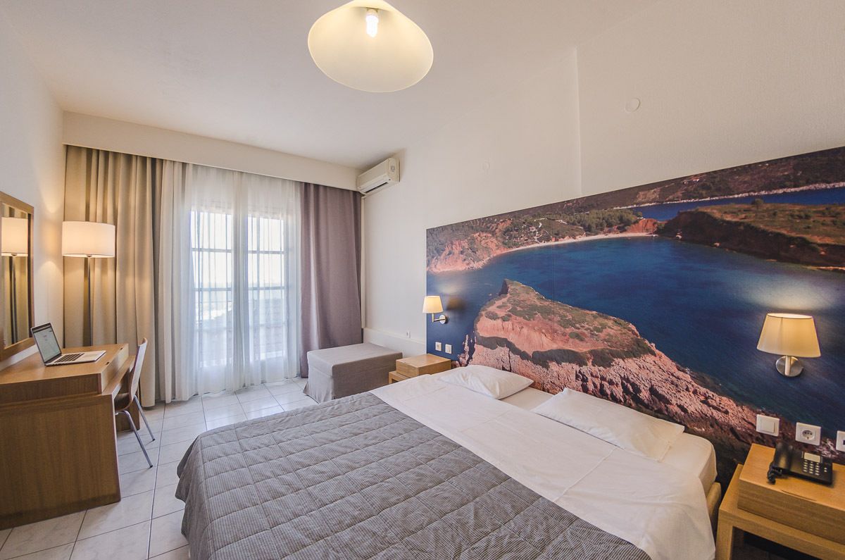 A hotel room with a bed, desk, chair, and a large painting of a coastal landscape.