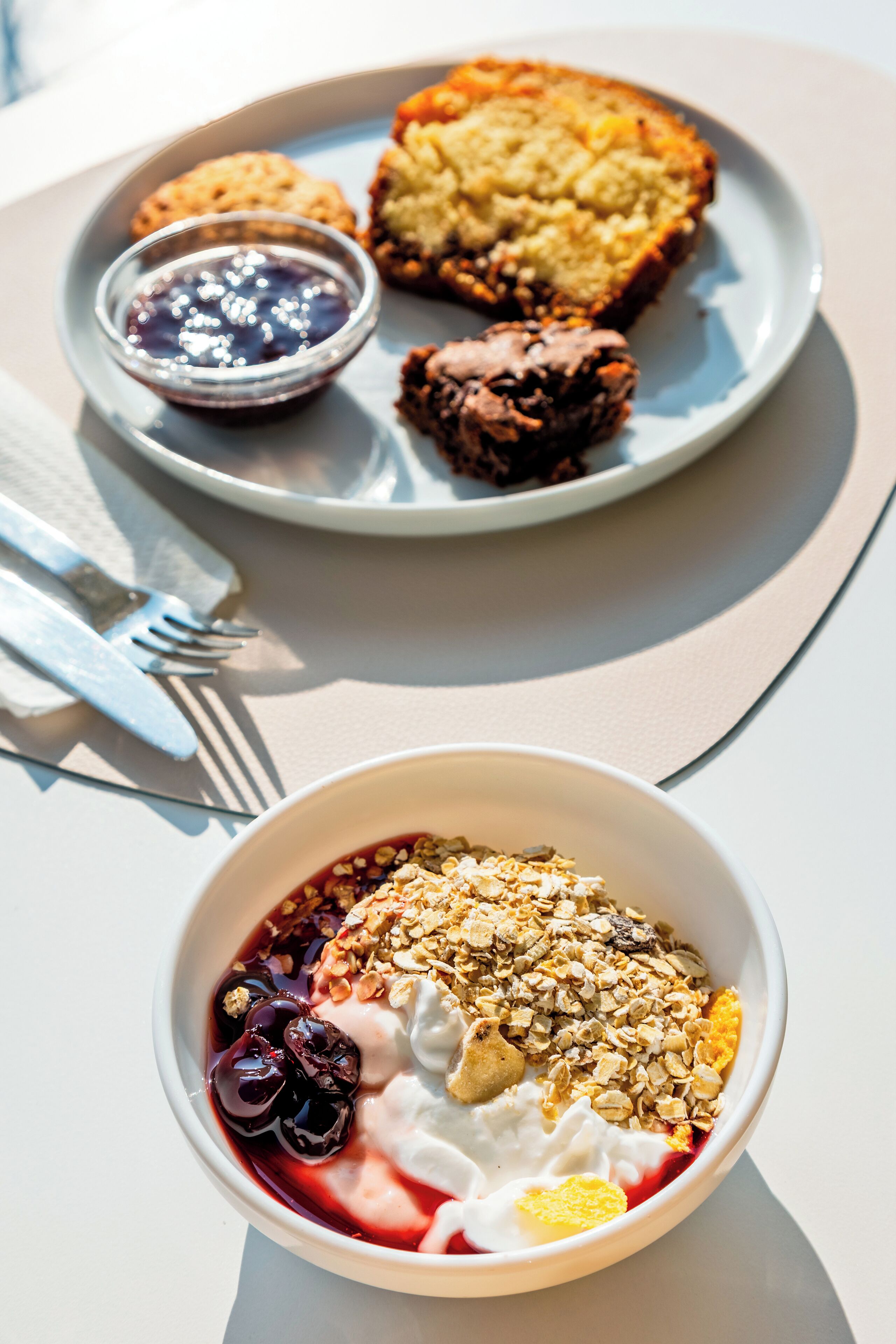 Una escena de desayuno con un plato de muffins y un tazón de yogur, granola y frutos rojos.