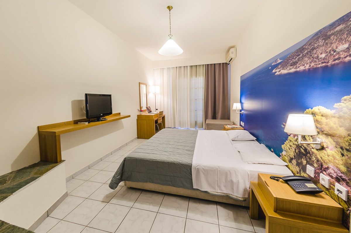 A hotel room with a bed, a desk with a computer, a TV, and a large window with curtains.