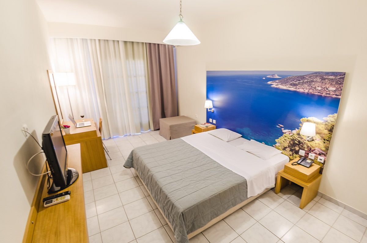 Une chambre d’hôtel équipée d’un lit, d’un bureau avec un ordinateur, d’une chaise, d’une télévision, d’une lampe et d’une grande fenêtre donnant sur la mer.
