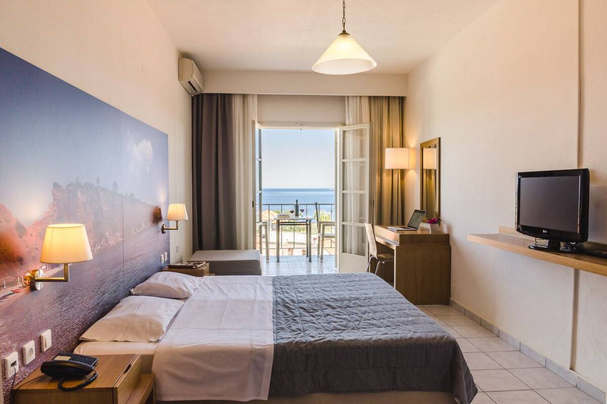 Una habitación de hotel con cama, lámparas de noche, un escritorio con computadora, televisión y un ventanal con vista al mar.