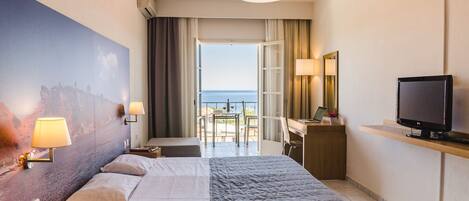 Una habitación de hotel con cama, lámparas de noche, un escritorio con computadora, televisión y un ventanal con vista al mar.