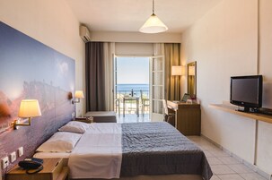 Una habitación de hotel con cama, lámparas de noche, un escritorio con computadora, televisión y un ventanal con vista al mar.