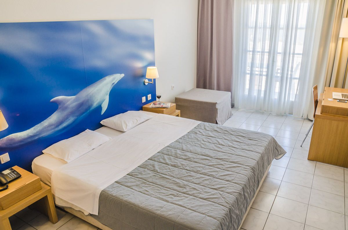 A hotel room with a large bed, a nightstand with a lamp, a wooden desk, and a window with curtains.