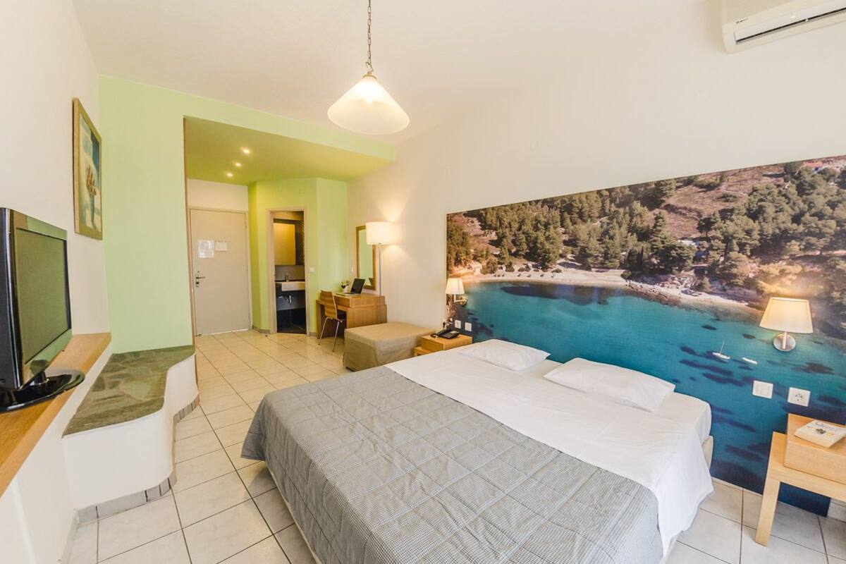 Habitación de hotel con cama, escritorio con computadora, silla, lámpara de escritorio y un cuadro de una playa.