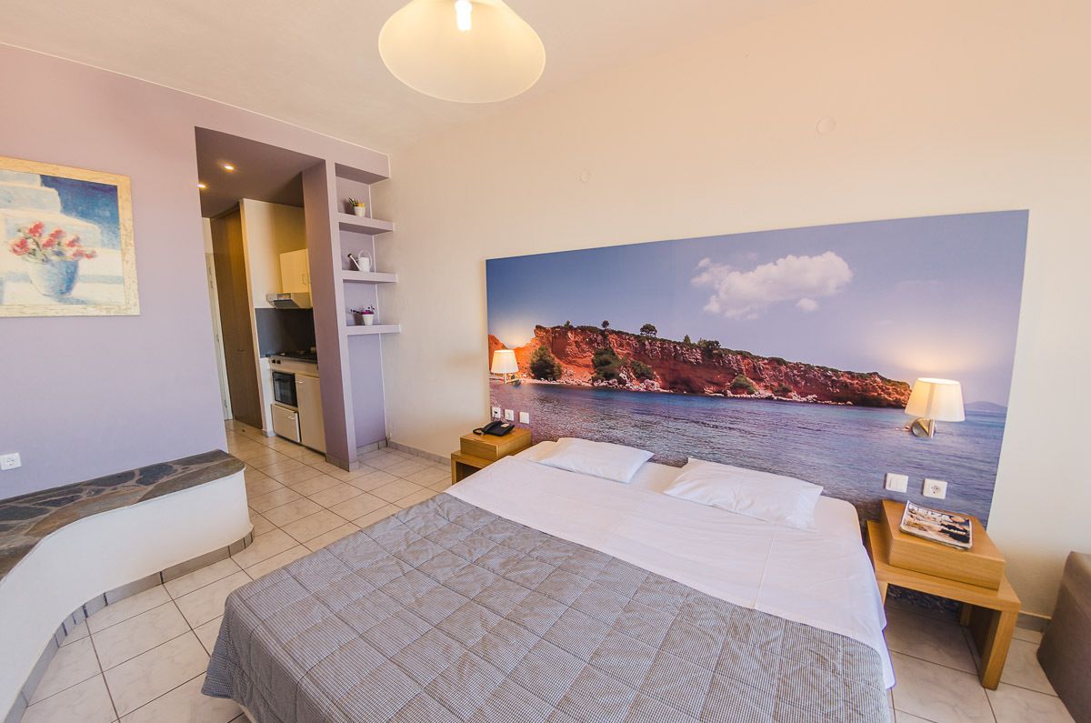 A hotel room with a bed, bedside tables, a nightstand, a painting of a coastal landscape, and a kitchenette in the background.