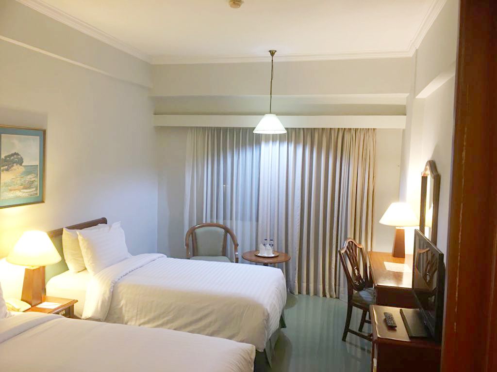 A hotel room with two beds, a desk, a chair, a lamp, a painting, and a curtain.