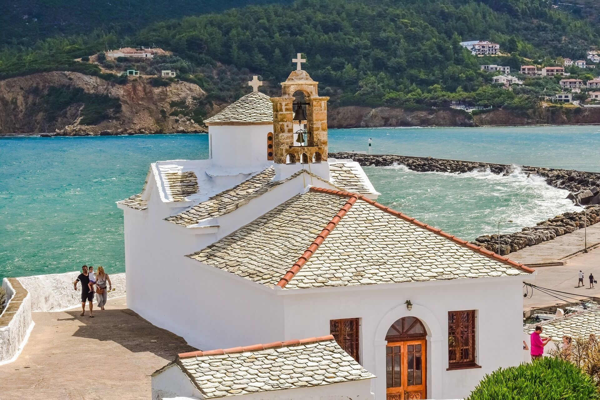 A coastal church with a white facade and a bell tower, situated near a rocky shoreline with a view of the sea and hills in the background.