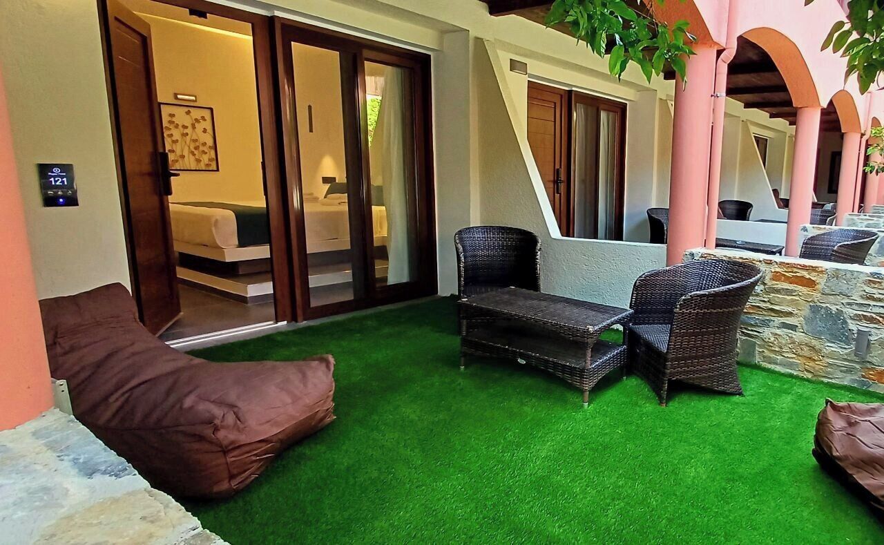 Una habitación con suelo de césped artificial verde, muebles de mimbre y una puerta corrediza de vidrio que da a un dormitorio.