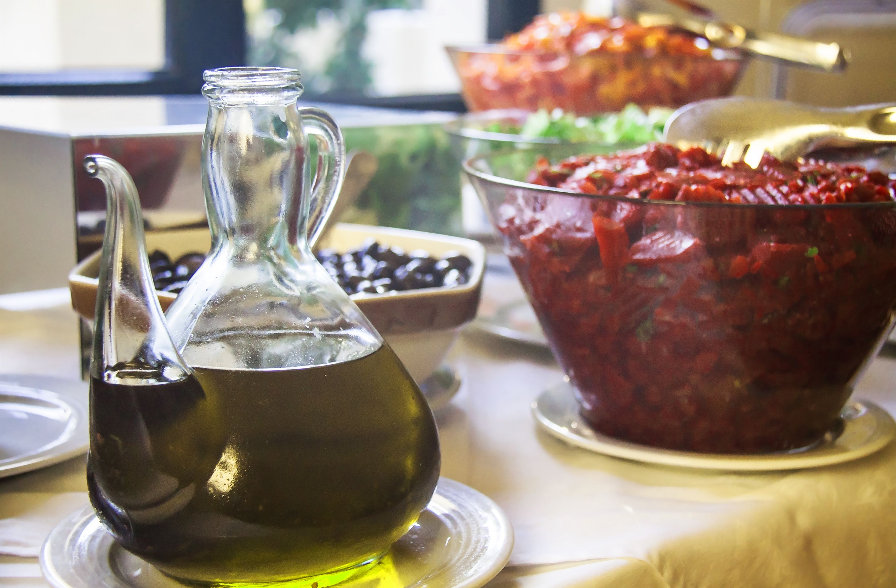 Una mesa con una botella de vidrio de aceite de oliva, un bol de verduras rojas y un bol de aceitunas.