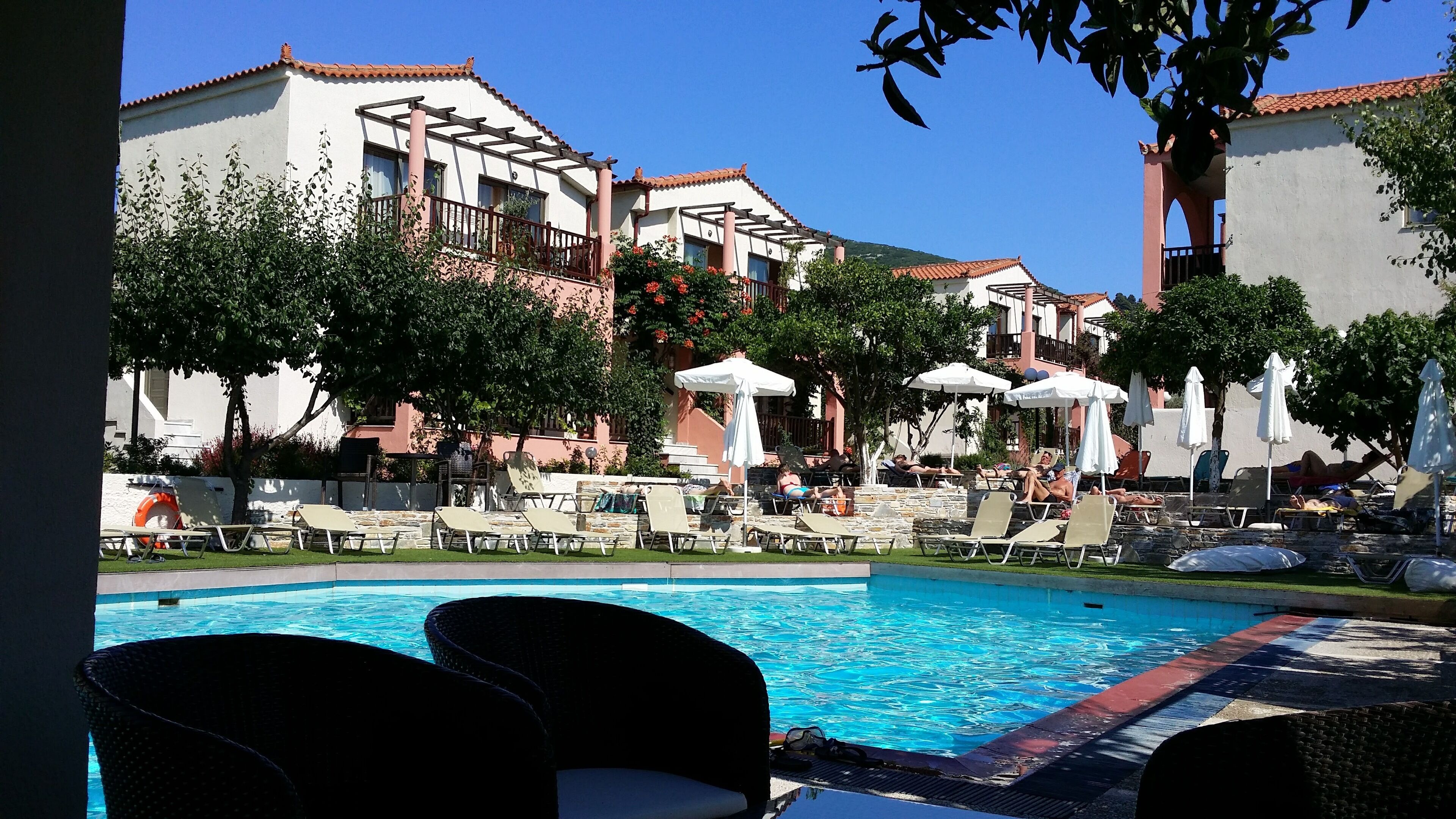 Un espace aménagé au bord de la piscine, avec des chaises longues et des parasols, entouré de bâtiments aux toits recouverts de tuiles rouges et dotés de balcons.