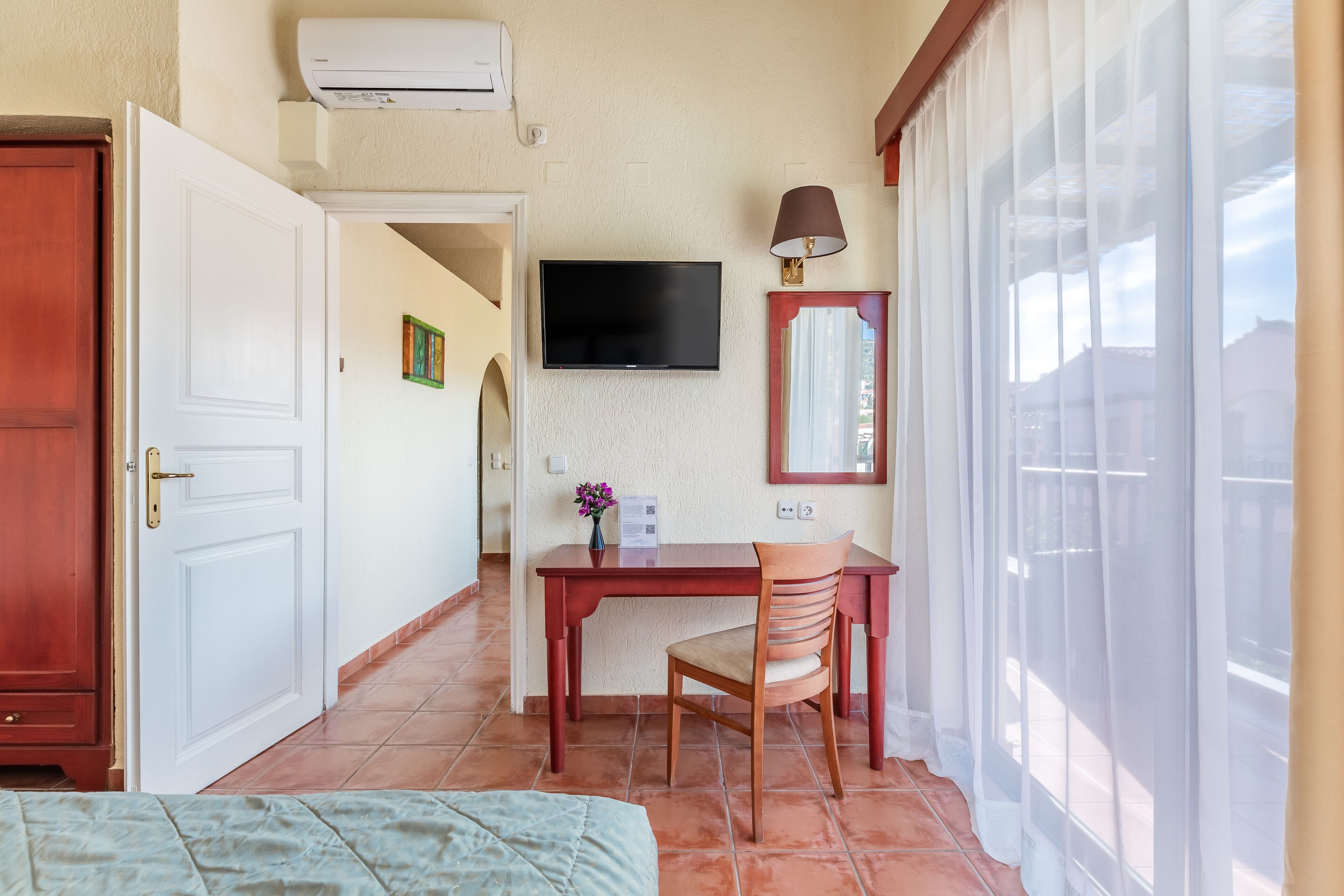A hotel room with a bed, a desk with a chair, a TV mounted on the wall, and a window with curtains.