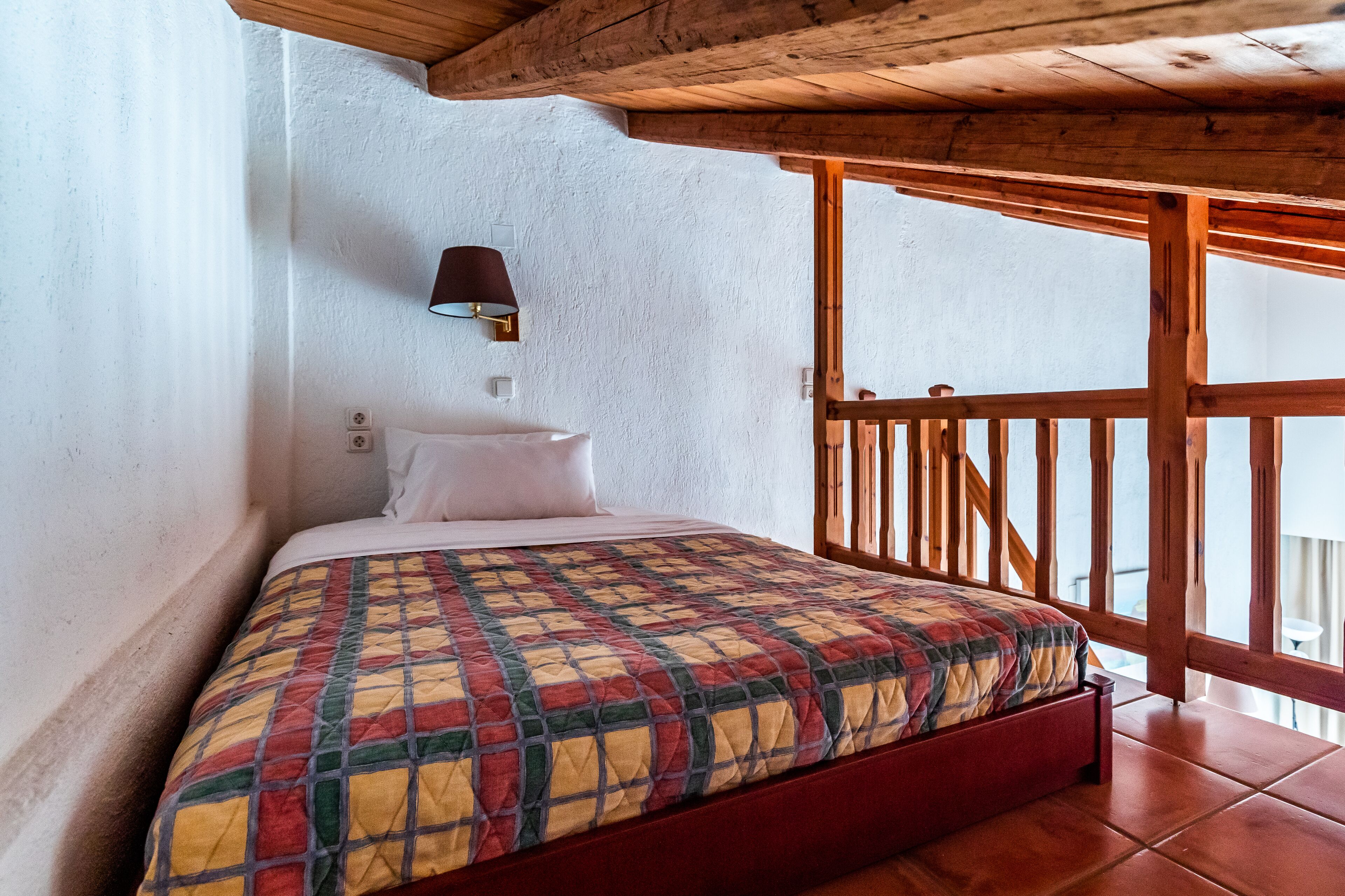 A bedroom with a wooden ceiling, a bed with a colorful quilt, a nightstand, and a wall-mounted lamp.