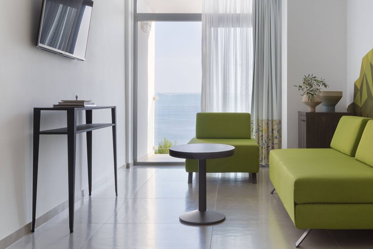 A modern living room with a green sofa, a round table, and a view of the sea through large windows.