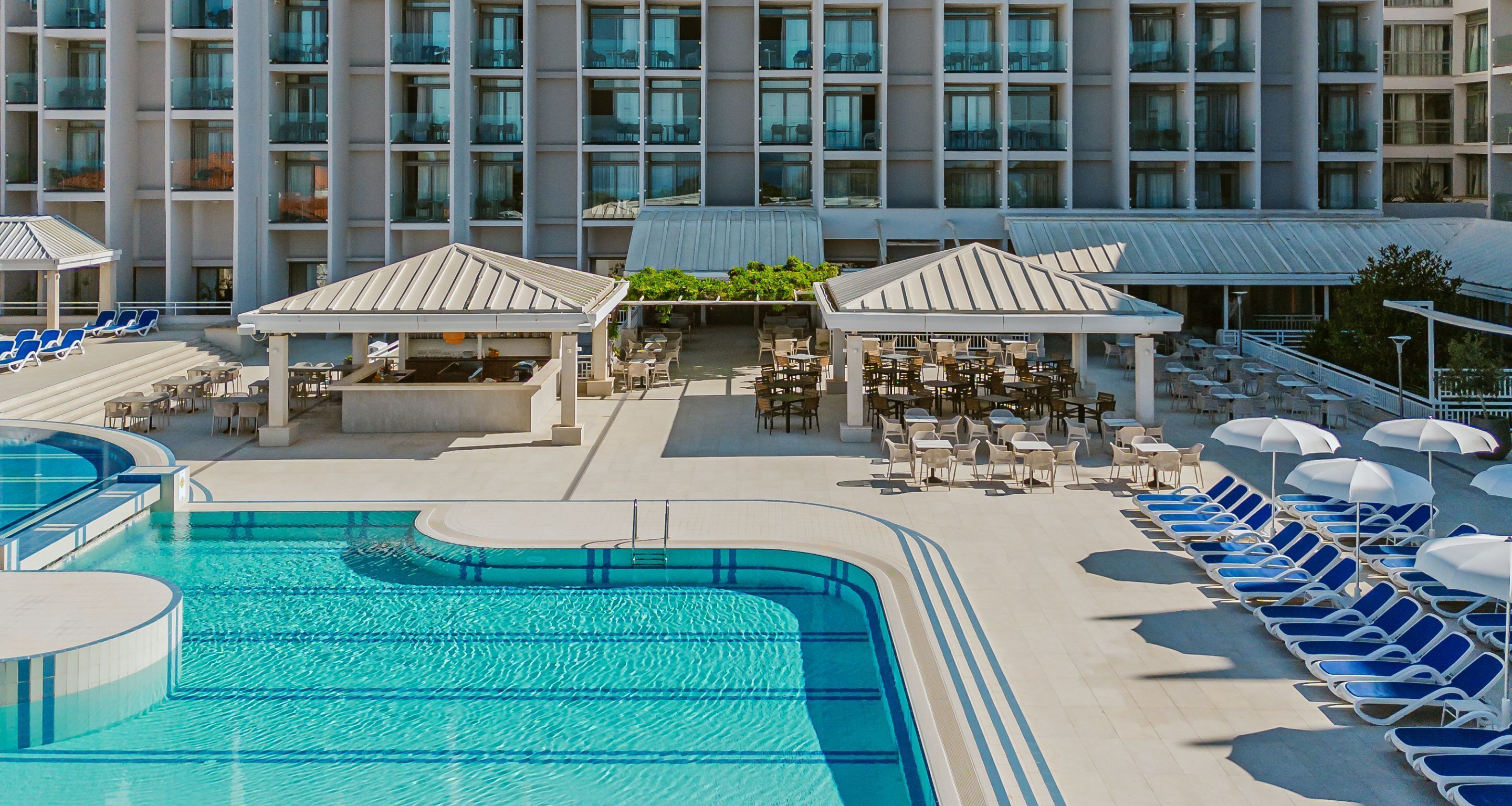Un área de piscina de hotel con múltiples sillas de descanso y sombrillas, una piscina y un edificio con balcones al fondo.
